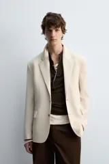 Blazer color beige confeccionado en tejido con lana, de corte relajado. Tiene cuello con solapas de muesca, manga larga con puños abotonados, bolsillo de vivo en el pecho, bolsillos de solapa en la cadera, bolsillo interior, bajo con abertura en el centro de la espalda y cierre frontal con botones.