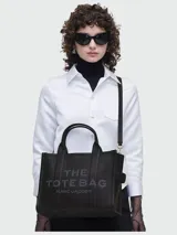 Bolso tote de cuero negro Marc Jacobs, tamaño mediano, con doble asa corta y correa larga extraíble. Estampado frontal con el texto "THE TOTE BAG" y el nombre de la marca.