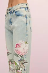 Pantalón de jean celeste con lavado vintage y calce mom. Presenta parches bordados con motivos florales, de mariposas y colibríes. El ruedo está desflecado.