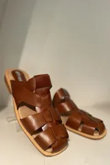 Zueco marrón de cuero vacuno con diseño de tiras entrecruzadas y suela plana.