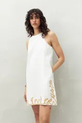 Vestido corto blanco de algodón con bordado artesanal color dorado en el ruedo. Tiene cuello alto y silueta recta.