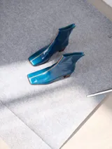 Botas de cuero charol negro con caña media y punta cuadrada. Cuentan con cierre trasero, tirador y bordado contrastante en el empeine. El taco es de madera y la suela de goma.