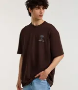 Remera marrón de corte relajado, confeccionada en algodón. Presenta cuello redondo, mangas cortas y estampa frontal y trasera de palmeras con la leyenda 'Work Hard'.