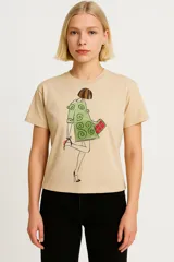Remera color beige de algodón con cuello redondo y mangas cortas. Estampado frontal con ilustración de una mujer con vestido verde, zapatos rojos y cartera roja.