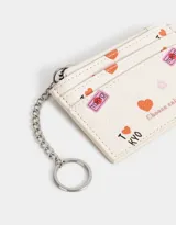 Tarjetero de cuerina color crema con estampado de corazones, flores y textos alusivos a Tokio y Kioto. Tiene cierre superior y mosquetón metálico plateado.