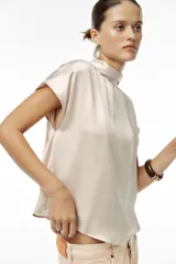 Blusa color crudo de tejido satinado, con cuello subido, manga sisa y cierre en la espalda con lazada y botones.