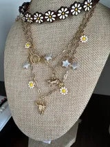 Collar dorado con cadena de eslabones y dijes colgantes: estrellas blancas, margaritas blancas y cabeza de toro dorada.