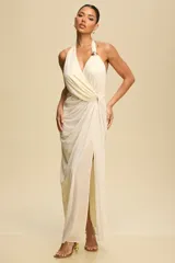 Vestido largo de fiesta color crema, con escote halter y espalda descubierta. Presenta un diseño drapeado con abertura en la falda y detalle de aro dorado en el cuello.