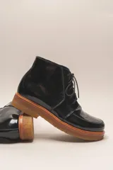 Bota de cuero vacuno con acabado charol, color marrón oscuro. Presenta un diseño de caña corta con cordones frontales y costuras decorativas en la puntera. La suela es de goma tipo crepe.