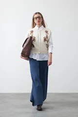 Chaleco de punto color blanco con bordado aplicado de cordón marrón en diseño floral. Presenta cierre frontal de botones al tono, cuello redondo y terminaciones acanaladas en sisas y ruedo.