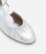 Zapatilla metalizada plateada con lazo para atar en la pierna, puntera redonda y detalle fruncido en la parte superior.