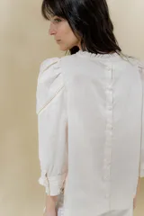 Blusa de lino blanca con escote a la base y terminación de voladito al bies desflecado. Delantera con alforzas verticales y mangas románticas tres cuartos, con detalle de puntilla. Puños con alforzas y voladitos que suman un toque femenino. Espalda abotonada.