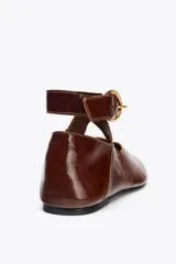 Zapato tipo bailarina color marrón, con efecto acharolado, punta redonda y cierre mediante tira con hebilla al tobillo.