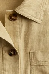 Campera color beige de sarga de algodón, con cuello, botones al frente y canesú en la espalda. Tiene hombros caídos, bolsillos superiores de parche, bolsillos insertados al frente y puños abotonados.