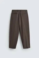 Pantalón negro de corte ancho con cintura elástica, pliegues frontales, bolsillos laterales y traseros de vivo. Tiene detalle de hebilla metálica en la trabilla y cierre frontal con cremallera y botón.