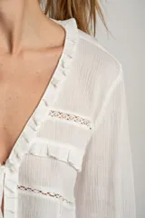 Blusa blanca de algodón con escote en V, cierre con lazos, mangas largas con puños elásticos y detalles de encaje y volados.