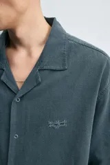 Camisa de denim de algodón, color beige claro, con corte relajado, cuello bowling y manga corta. Presenta cierre frontal con botones a presión y un pequeño bordado a tono en el pecho.