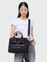 Bolso tote negro de lona con múltiples bolsillos externos, doble asa de mano y correa bandolera ajustable y extraíble. Estampado frontal con el texto "THE TOTE BAG MARC JACOBS".