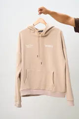 Conjunto deportivo color beige compuesto por un canguro con capucha y un pantalón jogging. El canguro tiene un bolsillo tipo canguro y estampados en el frente. El pantalón es de corte recto con cintura ajustable con cordón.