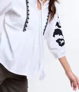 Blusa amplia de viscolino color blanco con bordados florales negros en mangas y escote, lazo en el cuello y mangas 3/4 con puño ajustado.