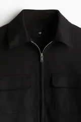Campera camisera negra de corte recto, con cuello clásico, cierre frontal con cremallera, dos bolsillos de parche con solapa en el pecho y bolsillos laterales.