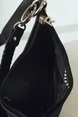 Cartera de hombro negra con forma de media luna, confeccionada en material con textura de pelo o cuero. Presenta costura decorativa gruesa en el borde y correa ajustable con herrajes metálicos plateados.
