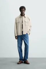 Sobrecamisa color beige de corte relaxed fit, confeccionada en tejido con elasticidad. Tiene cuello solapa, manga larga con puños abotonados, bolsillos de parche con solapa en el pecho y bolsillos de ojal en la cadera. El cierre es frontal con botones.