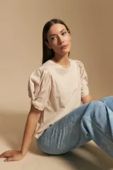 Remera de algodón color beige claro, con cuello redondo y mangas cortas abullonadas con detalle de frunce y ajuste en el puño.