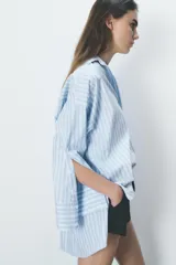 Camisa celeste y blanca a rayas verticales, de corte amplio, con cuello solapa, escote pico, manga larga con puños abotonados, bolsillo en el pecho y bajo asimétrico con aberturas laterales. Cierre frontal con botones.