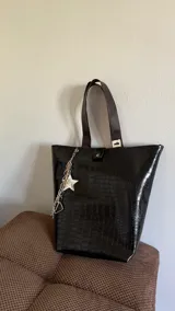 Bolso tote negro con textura símil cuero de cocodrilo, doble asa de hombro marrón y cierre con botón a presión. Incluye un charm colgante con una estrella blanca y cadenas plateadas.