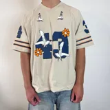 Remera color crema de corte oversize, con cuello en V y mangas cortas. Estampado frontal con el número 43 en azul, flores naranjas y gaviotas blancas. Tiene rayas azules y naranjas en las mangas.