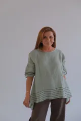 Blusa verde claro de lino, corte holgado, con cuello redondo y mangas 3/4. Presenta un diseño asimétrico con terminación en puntas y detalle de líneas horizontales negras en el ruedo.