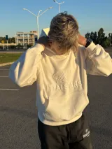 Buzo hoodie color crema con bolsillo tipo canguro y logo de la marca bordado en el frente.