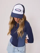 Gorra de lona blanca con visera azul marino y malla trasera azul marino. Presenta un logo bordado en el frente con letras rosadas y contorno azul marino.
