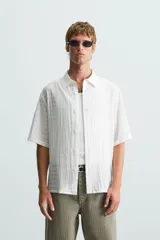 Camisa de hombre de manga corta, cuello solapa y corte relaxed fit. Tejido blanco con textura sutilmente arrugada o plisada. Cierre frontal de botonadura y bajo con aberturas laterales.