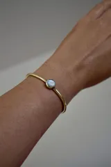 Pulsera rígida dorada con una piedra redonda de nácar en el centro.