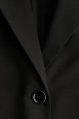 Blazer largo negro, de corte recto y botonadura sencilla con dos botones frontales. Presenta solapas de muesca, bolsillos delanteros ribeteados con solapa y hombreras.