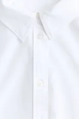 Camisa blanca de algodón con cuello clásico, abotonada adelante y corte holgado. Tiene mangas largas con puños sencillos y bajo redondeado con espalda algo más larga.