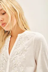 Camisa blanca de manga larga con cuello redondo y bordado floral en el delantero. El delantero es de algodón plano, mientras que las mangas y la espalda son de jersey a tono. Presenta un efecto fruncido en el centro de la espalda y puños en las mangas largas.
