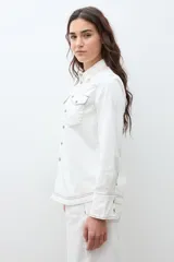 Camisa de jean color crudo, estilo western, con cuello y dos bolsillos frontales con tapa y botón. Presenta costuras contrastantes en tono rojizo.