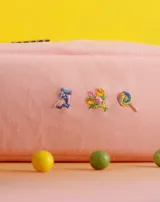 Neceser naranja de tela con cierre superior y logo de la marca en etiqueta negra. Presenta bordados de emojis: copa de vino, fuegos artificiales y mano verde.