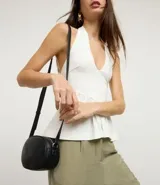 Cartera tipo camera bag pequeña, color marrón camel, de forma ovalada con superficie texturizada. Cuenta con correa ajustable larga, cierre principal con cremallera dorada y un pequeño bolsillo frontal con cremallera.