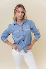 Camisa de denim celeste lavado, con cuello clásico y cierre de botones. Presenta bordado blanco en los bordes superiores de los bolsillos frontales y en los puños.