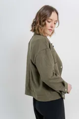 Chaqueta color beige de corte recto, estilo camisa, confeccionada en gamuza con elastano. Presenta cuello camisero, cierre frontal con botones metálicos, bolsillos grandes en el frente y charreteras en los hombros.