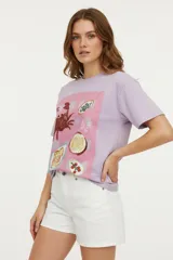 Remera de algodón color lila, de manga corta y cuello redondo. Presenta un estampado frontal con un cangrejo y diferentes platos de comida sobre un fondo rosado a cuadros. Algunos elementos del estampado tienen detalles bordados.