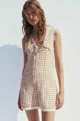 Vestido corto de escote pico con cuello bobo y manga sisa. Detalle de flores bordadas y entredoses. Forro interior. Cierre frontal con botones forrados y lazada. Estampado a cuadros vichy color marrón y blanco.