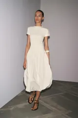 Vestido largo color crema, con cuello redondo y mangas cortas. Presenta corte a la altura de la cintura y falda amplia con terminación redondeada tipo globo.