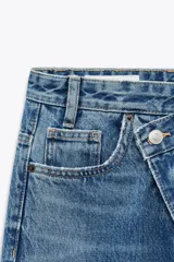 Pantalón de jean celeste de tiro alto, corte wide leg y largo hasta el tobillo. Presenta un diseño asimétrico en la cintura con cierre frontal cruzado con cremallera y botones metálicos.