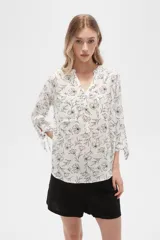 Blusa color beige con estampado de arabescos blancos, cuello mao, mangas 3/4 con lazos en los puños, cierre parcial con botones y bolsillos frontales.