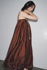 Vestido largo marrón de tejido satinado, con escote recto, tirantes finos extraíbles, bolsillos laterales ocultos y bajo asimétrico acabado en globo.